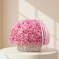 A large round arrangement of pink roses in a white wicker basket, tied with a pale pink ribbon, set on a marble surface in a softly lit indoor room.