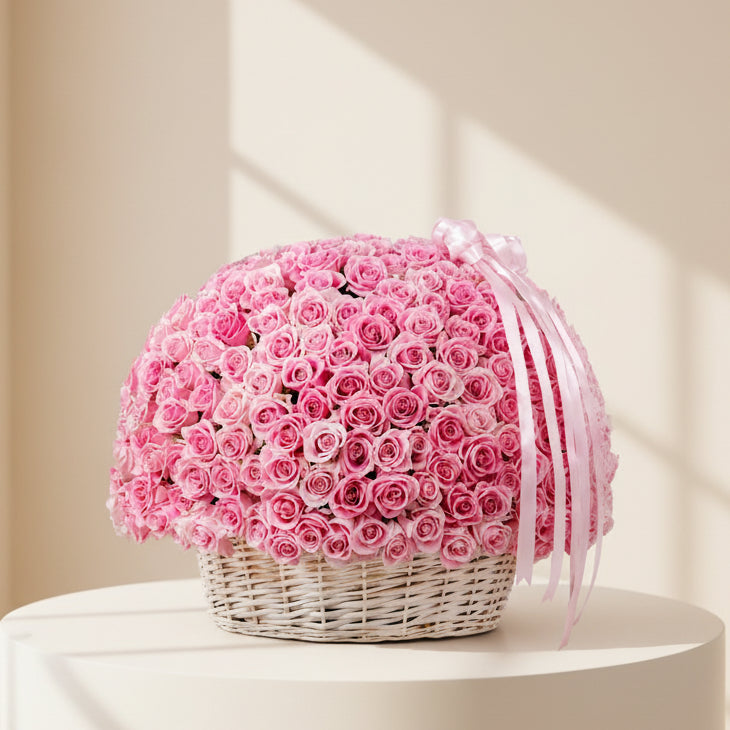 A large round arrangement of pink roses in a white wicker basket, tied with a pale pink ribbon, set on a marble surface in a softly lit indoor room.