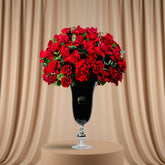 A large bouquet of vibrant red roses with green leaves arranged in a tall black glass vase with a silver pedestal base, set on a white surface against a plain light gray background.