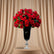 A large bouquet of vibrant red roses with green leaves arranged in a tall black glass vase with a silver pedestal base, set on a white surface against a plain light gray background.