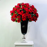 A large bouquet of vibrant red roses with green leaves arranged in a tall black glass vase with a silver pedestal base, set on a white surface against a plain light gray background.