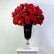 A large bouquet of vibrant red roses with green leaves arranged in a tall black glass vase with a silver pedestal base, set on a white surface against a plain light gray background.