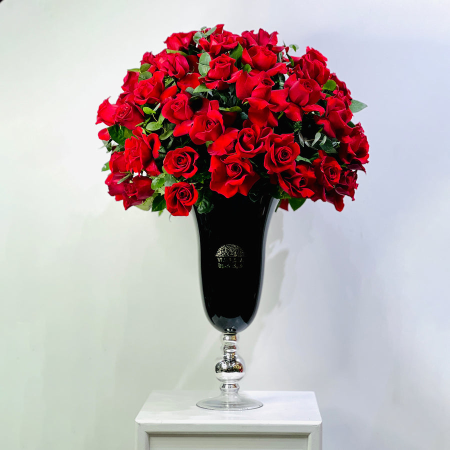 A large bouquet of vibrant red roses with green leaves arranged in a tall black glass vase with a silver pedestal base, set on a white surface against a plain light gray background.