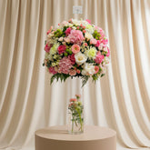 A large round floral arrangement in a tall clear glass vase, featuring pink, peach, cream, and white flowers with green foliage, displayed indoors against a light wall.
