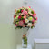 A large round floral arrangement in a tall clear glass vase, featuring pink, peach, cream, and white flowers with green foliage, displayed indoors against a light wall.