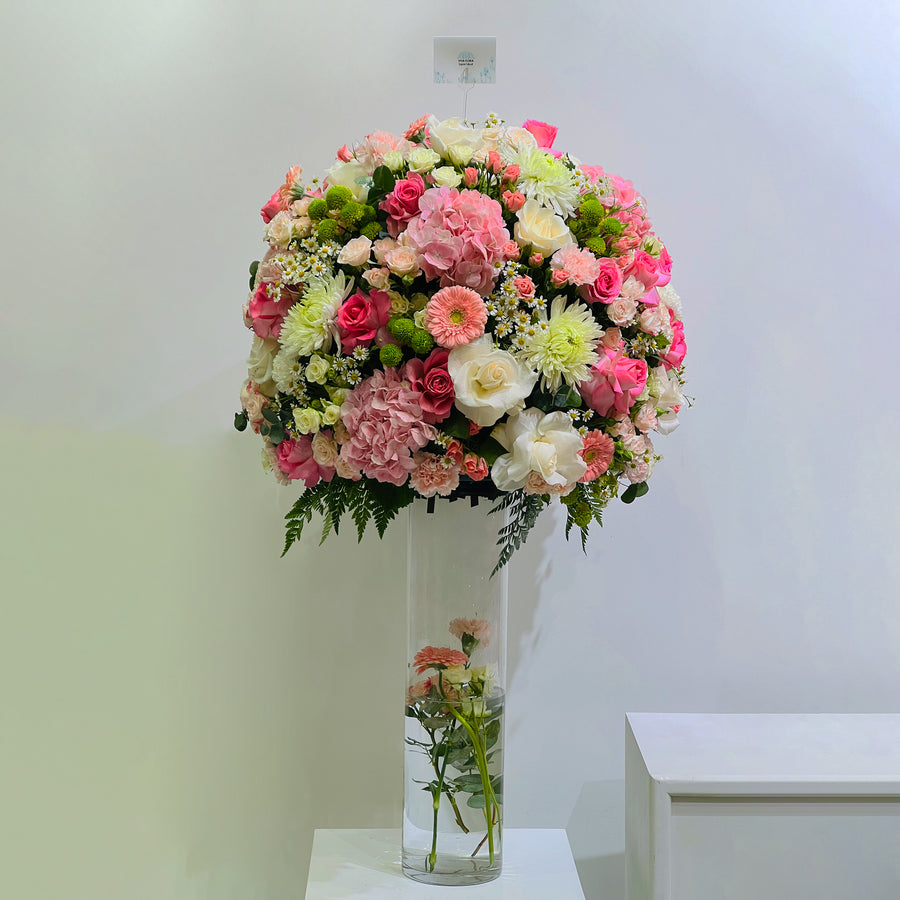 A large round floral arrangement in a tall clear glass vase, featuring pink, peach, cream, and white flowers with green foliage, displayed indoors against a light wall.