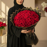A large bouquet of red roses wrapped in black and gold tissue paper, resting on a white pedestal against a beige wall with soft window shadows.