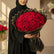 A large bouquet of red roses wrapped in black and gold tissue paper, resting on a white pedestal against a beige wall with soft window shadows.