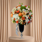 A large, vibrant artificial flower arrangement in a tall black pedestal vase with a silver base, set against a light gray wall with soft palm leaf shadows.