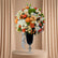 A large, vibrant artificial flower arrangement in a tall black pedestal vase with a silver base, set against a light gray wall with soft palm leaf shadows.
