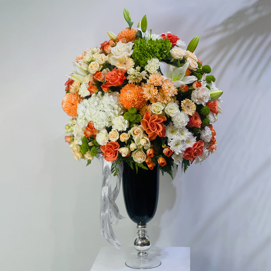 A large, vibrant artificial flower arrangement in a tall black pedestal vase with a silver base, set against a light gray wall with soft palm leaf shadows.