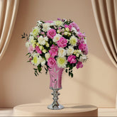 A large floral arrangement featuring pink and white roses, white and pink chrysanthemums, and green foliage, displayed in a pink vase with a silver pedestal base on a white surface against a plain light gray background.
