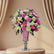A large floral arrangement featuring pink and white roses, white and pink chrysanthemums, and green foliage, displayed in a pink vase with a silver pedestal base on a white surface against a plain light gray background.
