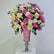 A large floral arrangement featuring pink and white roses, white and pink chrysanthemums, and green foliage, displayed in a pink vase with a silver pedestal base on a white surface against a plain light gray background.