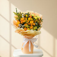 A hand holding a vibrant bouquet of yellow and orange roses with green foliage, wrapped in peach-colored paper and tied with a white ribbon, set against a blurred indoor background.