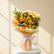 A hand holding a vibrant bouquet of yellow and orange roses with green foliage, wrapped in peach-colored paper and tied with a white ribbon, set against a blurred indoor background.