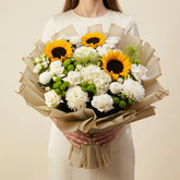 A large floral bouquet featuring sunflowers, white roses, white chrysanthemums, and green buds, wrapped in beige paper and displayed on a white pedestal against a white backdrop.