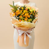 A vibrant bouquet of yellow and orange roses with green foliage, wrapped in peach-colored tissue paper and tied with a white ribbon, held by a person whose arm is visible on the left.
