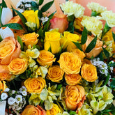 A vibrant bouquet of yellow and orange roses, yellow carnations, and green foliage wrapped in light peach paper.