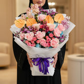 A vibrant floral bouquet wrapped in white paper and tied with a purple ribbon, featuring pink and orange flowers, set against a black square backdrop on a white marble surface.