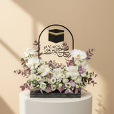 A person wearing a black abaya and headscarf holds a decorative black floral basket with a raised handle. The basket features a black cube design with gold trim at the top and Arabic calligraphy, filled with white and pale pink flowers and green foliage, against a warm beige background.