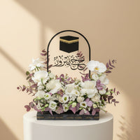 A person wearing a black abaya and headscarf holds a decorative black floral basket with a raised handle. The basket features a black cube design with gold trim at the top and Arabic calligraphy, filled with white and pale pink flowers and green foliage, against a warm beige background.
