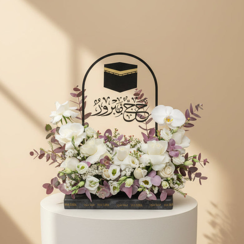 A person wearing a black abaya and headscarf holds a decorative black floral basket with a raised handle. The basket features a black cube design with gold trim at the top and Arabic calligraphy, filled with white and pale pink flowers and green foliage, against a warm beige background.