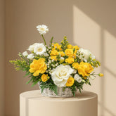 A person wearing a beige hijab holds a white wicker basket filled with a vibrant bouquet of yellow and white flowers, including roses, daisies, and green foliage, against a soft beige background.