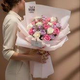 A bouquet of pink, white, and lavender roses wrapped in pale pink paper with a kraft ribbon, displayed on a white pedestal against a warm beige background.