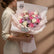 A bouquet of pink, white, and lavender roses wrapped in pale pink paper with a kraft ribbon, displayed on a white pedestal against a warm beige background.