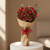 A person wearing a black abaya holds a large bouquet of red roses wrapped in brown paper with a red ribbon, set against a softly lit beige indoor background.