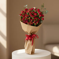 A person wearing a black abaya holds a large bouquet of red roses wrapped in brown paper with a red ribbon, set against a softly lit beige indoor background.