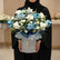 A floral arrangement in a round white box, featuring blue and white roses, baby's breath, and green foliage, with a blue ribbon bow and a small script card, displayed on a white pedestal against a warm beige background with soft window shadows.