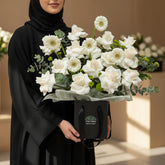 A floral arrangement of white roses, daisies, and green eucalyptus stems in a black gift bag with a circular logo, displayed on a white cylindrical pedestal against a warm beige wall with soft window-shadow lighting.