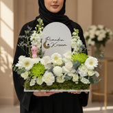 A floral arrangement on a round white pedestal against a warm beige wall with soft window-shadow lighting. The bouquet includes white and cream flowers, green foliage, and a white arched sign reading 'Ramadan Kareem' with a small gold crescent moon.