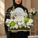 A floral arrangement on a round white pedestal against a warm beige wall with soft window-shadow lighting. The bouquet includes white and cream flowers, green foliage, and a white arched sign reading 'Ramadan Kareem' with a small gold crescent moon.