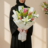 A wrapped bouquet of lilies and greenery on a white pedestal against a warm beige wall with soft window-shadow light. The flowers include white lilies, pink lilies, yellow buds, and green leaves, wrapped in white paper with gold trim and a pink ribbon.