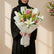 A wrapped bouquet of lilies and greenery on a white pedestal against a warm beige wall with soft window-shadow light. The flowers include white lilies, pink lilies, yellow buds, and green leaves, wrapped in white paper with gold trim and a pink ribbon.
