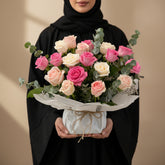 A bouquet of pink and cream roses with green eucalyptus stems, wrapped in white tissue paper and tied with a gold ribbon, displayed on a round white tray against a warm beige background with soft window shadows.