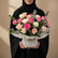 A bouquet of pink and cream roses with green eucalyptus stems, wrapped in white tissue paper and tied with a gold ribbon, displayed on a round white tray against a warm beige background with soft window shadows.