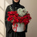 A heart-shaped flower arrangement featuring red roses, a central cluster of white baby's breath, and eucalyptus leaves, presented in a white cylindrical box with a red ribbon, set on a white pedestal against a beige background with soft window shadows.