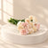 A small bouquet of carnation flowers with pale pink and cream petals and green stems, placed on a white surface against a white background.