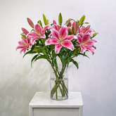 A bouquet of pink and white striped lilies in a clear glass vase, placed on a white side table against a light gray wall.