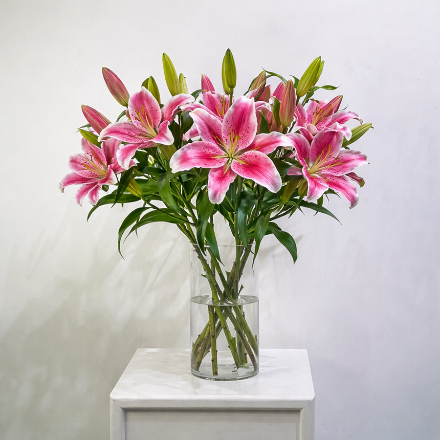 A bouquet of pink and white striped lilies in a clear glass vase, placed on a white side table against a light gray wall.