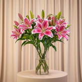 A bouquet of pink and white striped lilies in a clear glass vase, placed on a white side table against a light gray wall.