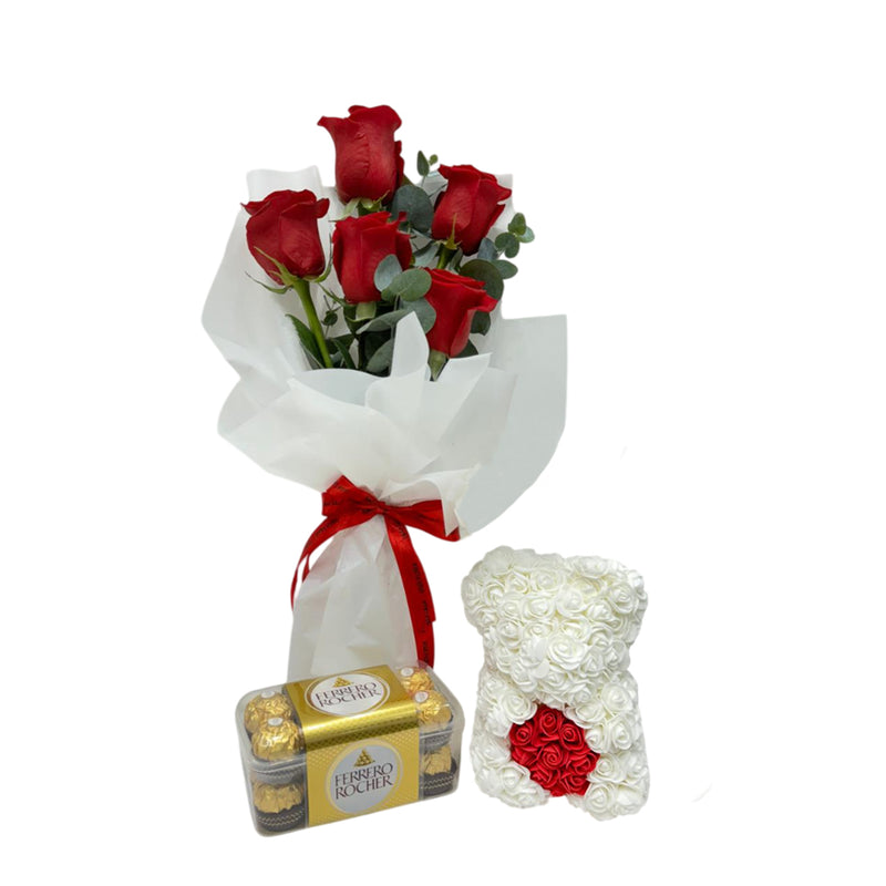 A bouquet of red roses wrapped in white paper with a red ribbon is placed next to a white floral teddy bear with a red rose heart accent.