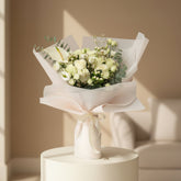 A person in a beige hijab holds a large bouquet of white and cream flowers wrapped in pale blush paper with a cream ribbon. The bouquet includes white roses, an allium, eucalyptus, and small filler flowers, with a small card attached.