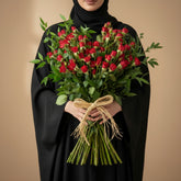 A vibrant bouquet of red roses with lush green leaves, tied with a natural jute ribbon, displayed on a white cylindrical pedestal against a neutral gray background.
