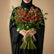 A vibrant bouquet of red roses with lush green leaves, tied with a natural jute ribbon, displayed on a white cylindrical pedestal against a neutral gray background.