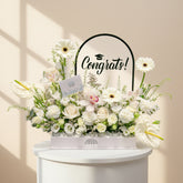 A person in a black abaya and headscarf holds a large white floral arrangement box with a clear graduation-themed sign reading 'Congrats!' behind it. The bouquet includes white and cream flowers, green foliage, and white anthuriums, with the brand name 'YAN FLORIST' visible on the box.